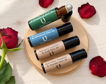May include: Four amber glass roll-on bottles with black caps, labeled Grapefruit, De-stress Bergamot & Geranium, Relaxing Lavender & Rose, and Motivating Patchouli & Jasmine. Each bottle features a U-shaped logo and is arranged on a round wooden disc.