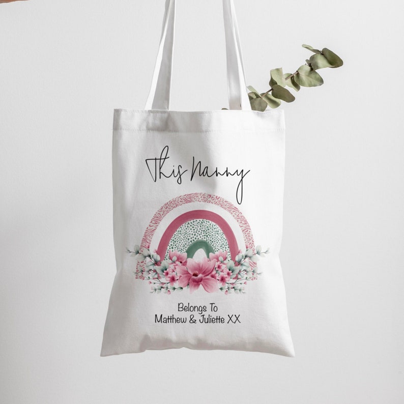 Op de afbeelding: Witte draagtas met de tekst "This Nanny" in zwart script. Een aquarel regenboogontwerp in roze, groen en wit is gecentreerd onder de tekst, met bloemaccenten en de tekst "Belongs To Matthew & Juliette XX".