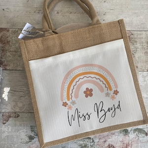 May include: A brown burlap tote bag with a white fabric insert. The insert features a rainbow with pink, orange, and yellow stripes and a scattering of small white flowers. The text "Miss Boyd" is written in black cursive below the rainbow.