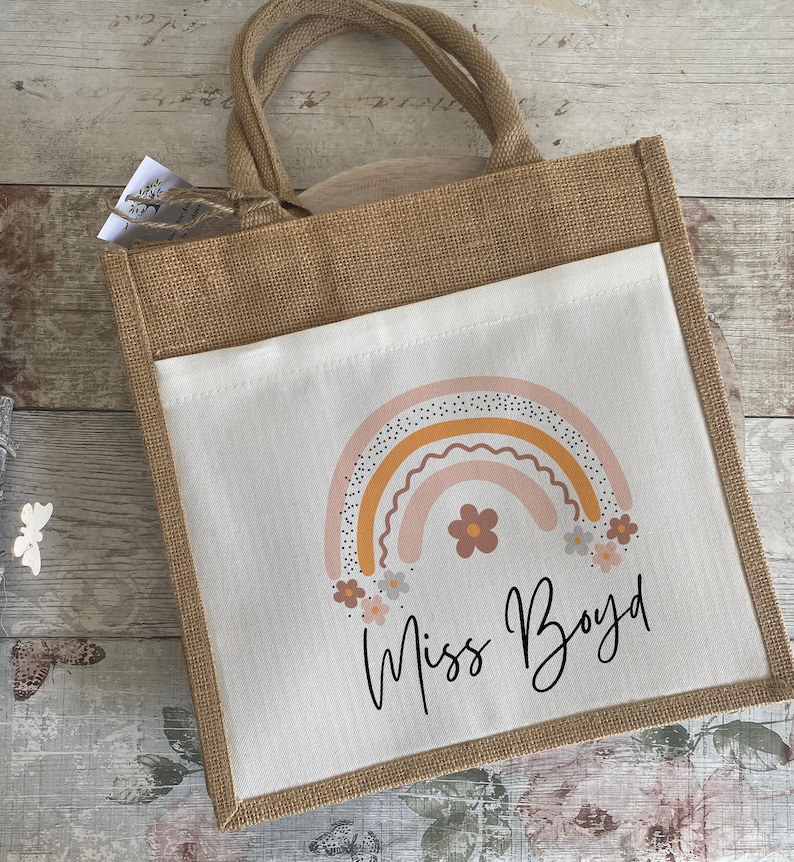 May include: A brown burlap tote bag with a white fabric insert. The insert features a rainbow with pink, orange, and yellow stripes and a scattering of small white flowers. The text "Miss Boyd" is written in black cursive below the rainbow.