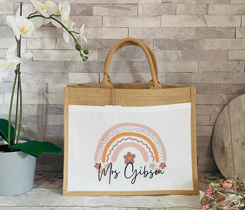May include: A brown burlap tote bag with a white insert featuring a rainbow design with pink, orange, and yellow colors. The text "Mrs Gibson" is printed below the rainbow.