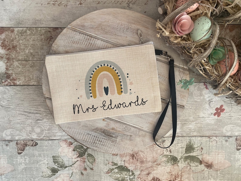 May include: A beige canvas zippered pouch with a rainbow design and the text "Mrs. Edwards". The rainbow is made of yellow, orange, pink, and blue stripes with black stars. The pouch has a black strap.