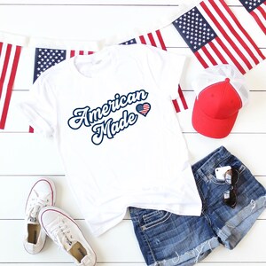 May include: White t-shirt with the text "American Made" in blue script with a red, white, and blue heart with a small American flag inside. The shirt is laying on a white wooden surface with blue denim shorts, a red baseball cap, and American flag bunting.