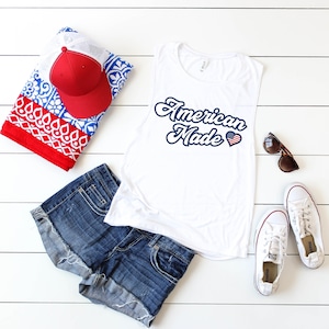 May include: White sleeveless muscle tank top with the text "American Made" in blue script with a red, white, and blue heart. The tank top is laying on a white wood surface with blue denim shorts, a red and white patterned beach towel, a red baseball cap, and white sneakers.