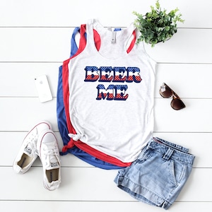May include: A white tank top with a red, white, and blue "BEER ME" graphic. The graphic is in a retro font and has a red, white, and blue star pattern. The tank top is laying on a white wooden surface with a pair of white sneakers, a pair of blue denim shorts, a white phone, and sunglasses.