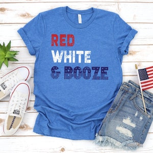 May include: Blue t-shirt with the text "RED WHITE & BOOZE" in red and white block letters. The word "BOOZE" is designed with blue and white stripes and stars. The shirt is paired with denim shorts, white sneakers, and a small American flag.