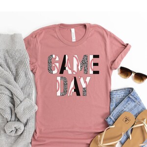 May include: A dusty rose t-shirt with the words "GAME DAY" printed on the front. The letters are made up of baseballs with a black and white leopard print pattern.