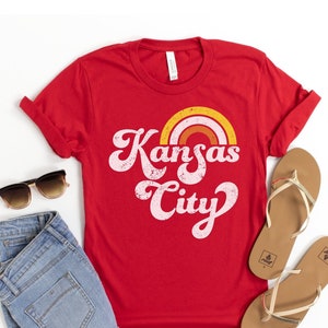 Op de afbeelding: Rood T-shirt met een retro-grafische afbeelding met de tekst "Kansas City" met een regenboog boven de tekst.