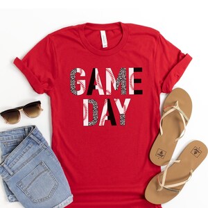 May include: Red t-shirt with the words "GAME DAY" in black letters with a baseball and leopard print design.