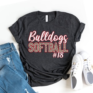 以下が含まれることがあります： ダークグレーのTシャツに、「Bulldogs Softball #18」という赤い文字と白い文字がレオパード柄で書かれています。