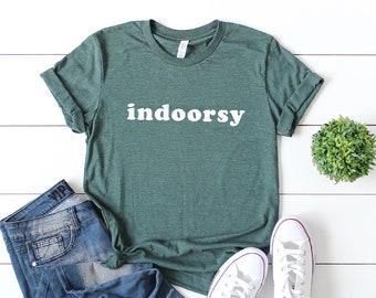 May include: A green t-shirt with the word "indoorsy" printed in white on the front. The shirt is paired with blue jeans and white sneakers.