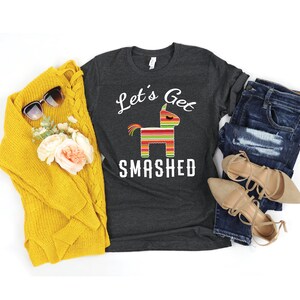 May include: A dark heather grey t-shirt with a white graphic of a pinata and the text "Let's Get Smashed".