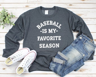 May include: A dark gray sweatshirt with the text "Baseball - Is My - Favorite Season" in white. The sweatshirt is laid flat on a wooden surface with a pair of white sneakers and blue jeans.