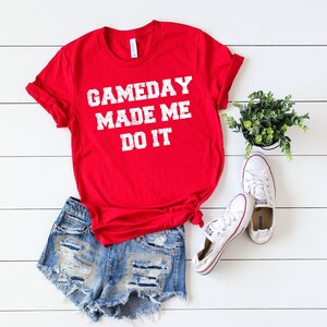 May include: Red t-shirt with white text that reads "GAMEDAY MADE ME DO IT". The shirt is paired with denim shorts and white sneakers.