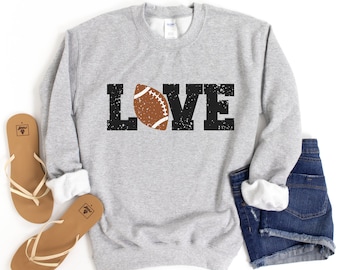 Puede incluir: Una sudadera gris con la palabra "LOVE" impresa en negro con una pelota de fútbol marrón en el medio de la palabra.