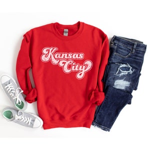 May include: Red sweatshirt with a white retro-style graphic that says "Kansas City". The sweatshirt is laid flat on a white surface with a pair of blue jeans and a pair of gray sneakers.