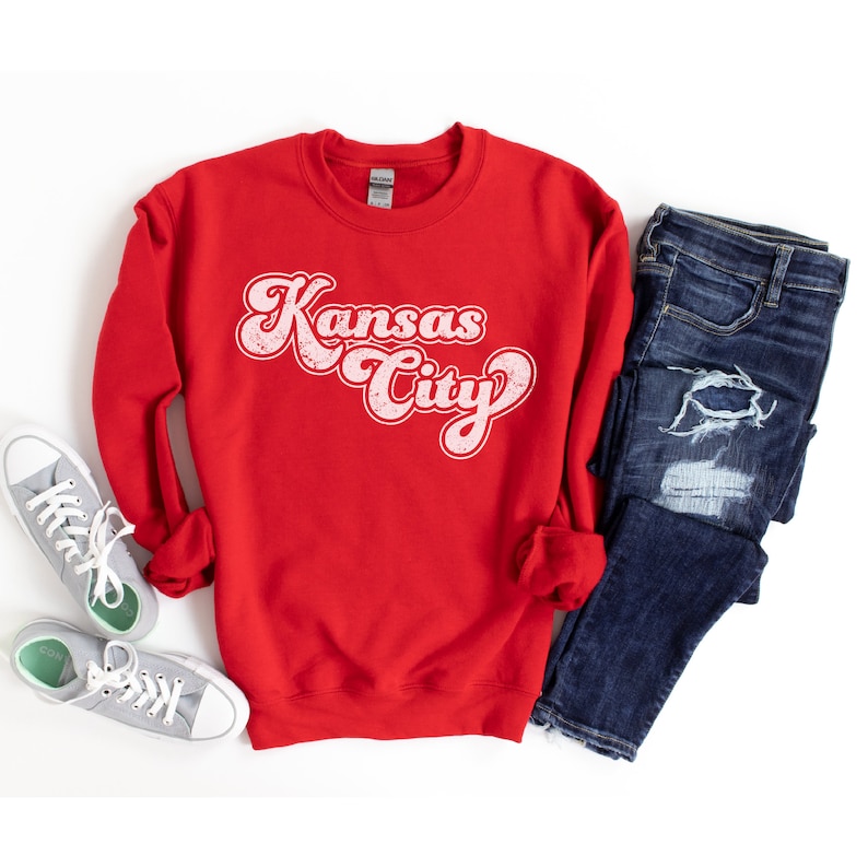 May include: Red sweatshirt with a white retro-style graphic that says "Kansas City". The sweatshirt is laid flat on a white surface with a pair of blue jeans and a pair of gray sneakers.