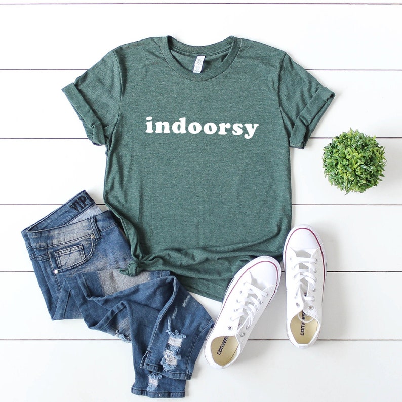 K&ouml;nnte beinhalten: Ein gr&uuml;nes T-Shirt mit dem Wort "indoorsy" in wei&szlig; auf der Vorderseite. Das Shirt wird mit einer blauen Jeans und wei&szlig;en Turnschuhen kombiniert.