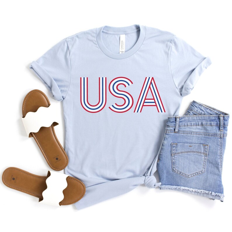 May include: A light blue t-shirt with the word "USA" printed in red and blue stripes. The shirt is paired with denim shorts and white sandals.