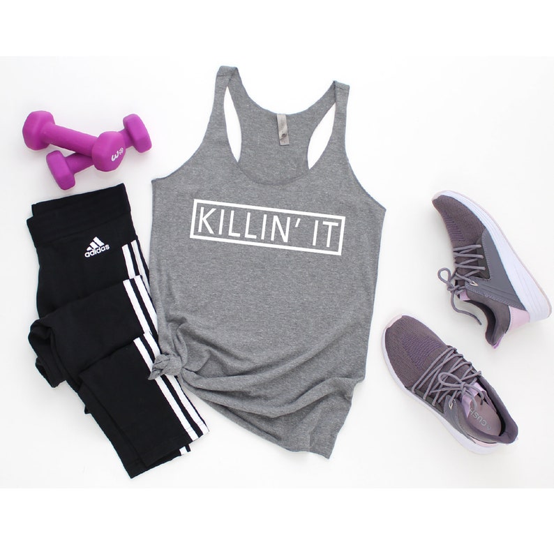 May include: Gray tank top with the text "KILLIN' IT" in a white box. The tank top is paired with black leggings with white stripes and purple dumbbells.