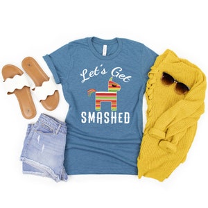 May include: A teal blue t-shirt with the text "Let's Get Smashed" and a colorful pinata graphic. The shirt is laid flat on a white background with a pair of white sandals, denim shorts, and a yellow sweater.