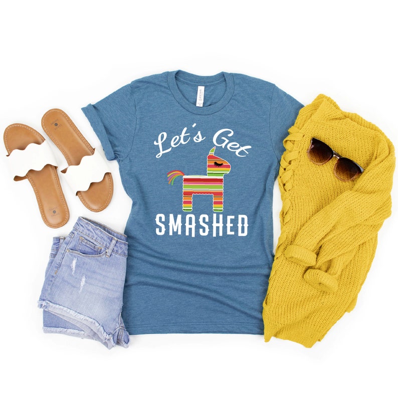 May include: A teal blue t-shirt with the text "Let's Get Smashed" and a colorful pinata graphic. The shirt is laid flat on a white background with a pair of white sandals, denim shorts, and a yellow sweater.