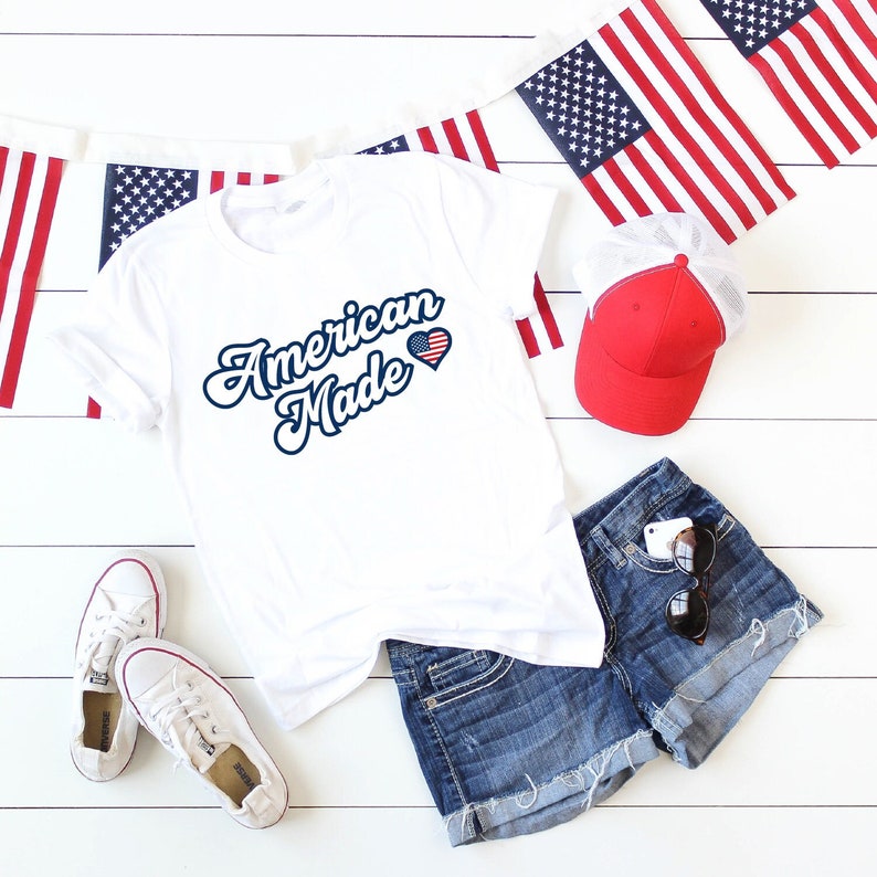 May include: White t-shirt with the text "American Made" in blue script with a red, white, and blue heart with a small American flag inside. The shirt is laying on a white wooden surface with blue denim shorts, a red baseball cap, and American flag bunting.