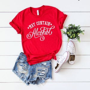 May include: Red t-shirt with the text "*MAY CONTAIN* Alcohol" in white cursive lettering. The shirt is paired with denim shorts and white sneakers.