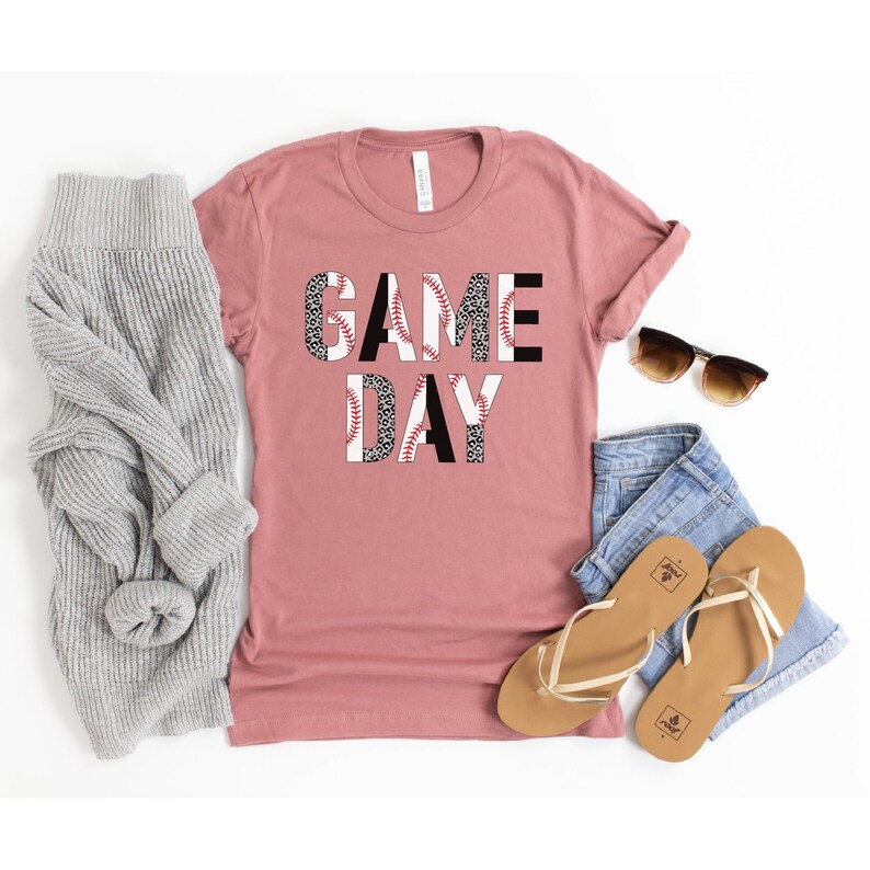 May include: A dusty rose t-shirt with the words "GAME DAY" printed on the front. The letters are made up of baseballs with a black and white leopard print pattern.
