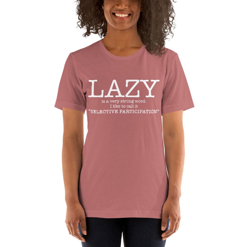 Op de afbeelding: Een roze T-shirt met witte tekst die "LAZY is a very strong word. I like to call it "SELECTIVE PARTICIPATION" zegt.