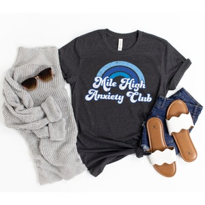 May include: A dark gray t-shirt with a blue and white rainbow graphic and the text "Mile High Anxiety Club". The shirt is shown on a white background with a gray sweater, sunglasses, and white sandals.