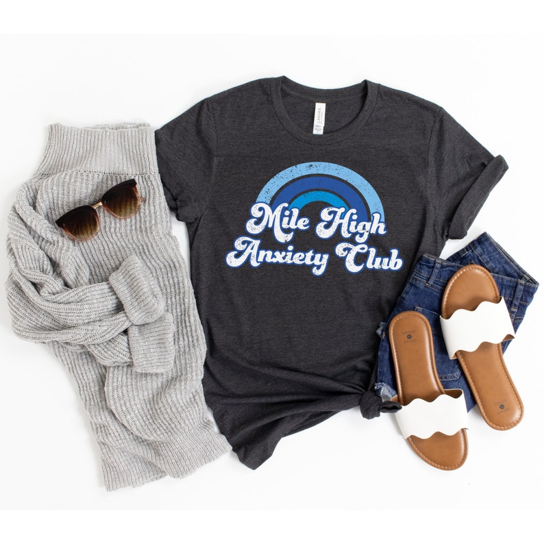 May include: A dark gray t-shirt with a blue and white rainbow graphic and the text "Mile High Anxiety Club". The shirt is shown on a white background with a gray sweater, sunglasses, and white sandals.