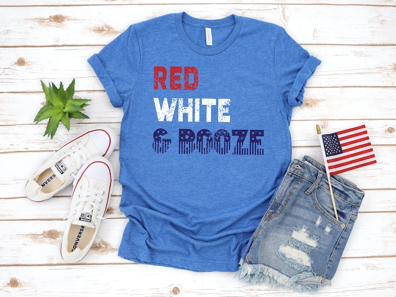 May include: Blue t-shirt with the text "RED WHITE & BOOZE" in red and white block letters. The word "BOOZE" is designed with blue and white stripes and stars. The shirt is paired with denim shorts, white sneakers, and a small American flag.