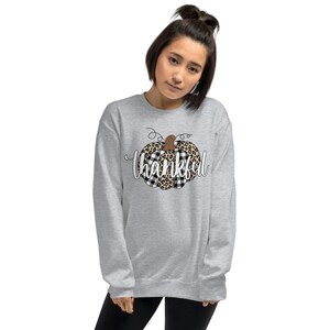 May include: A grey sweatshirt featuring a black and white checked and leopard print pumpkin design. The word "thankful" is written in white below the pumpkin.