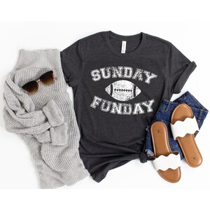 Op de afbeelding: Een donker grijs t-shirt met een witte afbeelding van een voetbal en de tekst "SUNDAY FUNDAY".