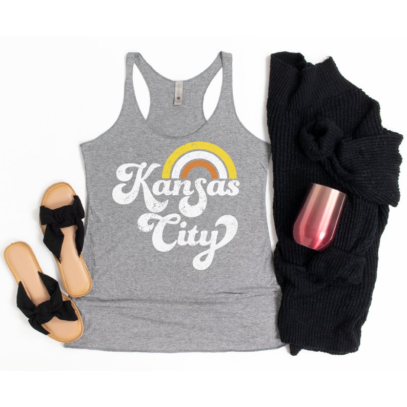Puede incluir: Un top de tirantes gris con un gr&aacute;fico de estilo retro que dice "Kansas City" con un arco&iacute;ris sobre el texto.