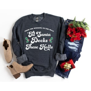 Può includere: Una felpa grigia scura con il testo "'Til Santa Decks These Halls" in bianco con accenti di agrifoglio verde. Il testo è in un font retrò. La felpa è stesa su uno sfondo bianco con un paio di stivaletti marroni alla caviglia, jeans blu e una scatola regalo rossa e nera.