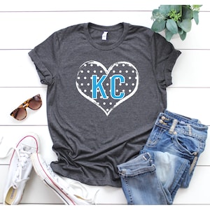 Pode incluir: Uma camiseta cinza escura com um design de cora&ccedil;&atilde;o branco com bolinhas azuis e as letras "KC" no centro.