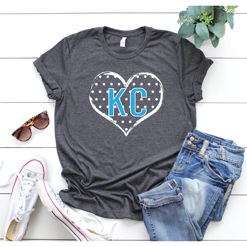 Pode incluir: Uma camiseta cinza escura com um design de cora&ccedil;&atilde;o branco com bolinhas azuis e as letras "KC" no centro.