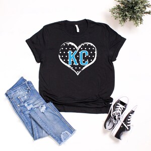 Pode incluir: Camiseta preta com um design de cora&ccedil;&atilde;o branco com bolinhas pretas e as letras "KC" em azul. A camiseta &eacute; exibida em uma superf&iacute;cie branca com jeans azul e t&ecirc;nis pretos.