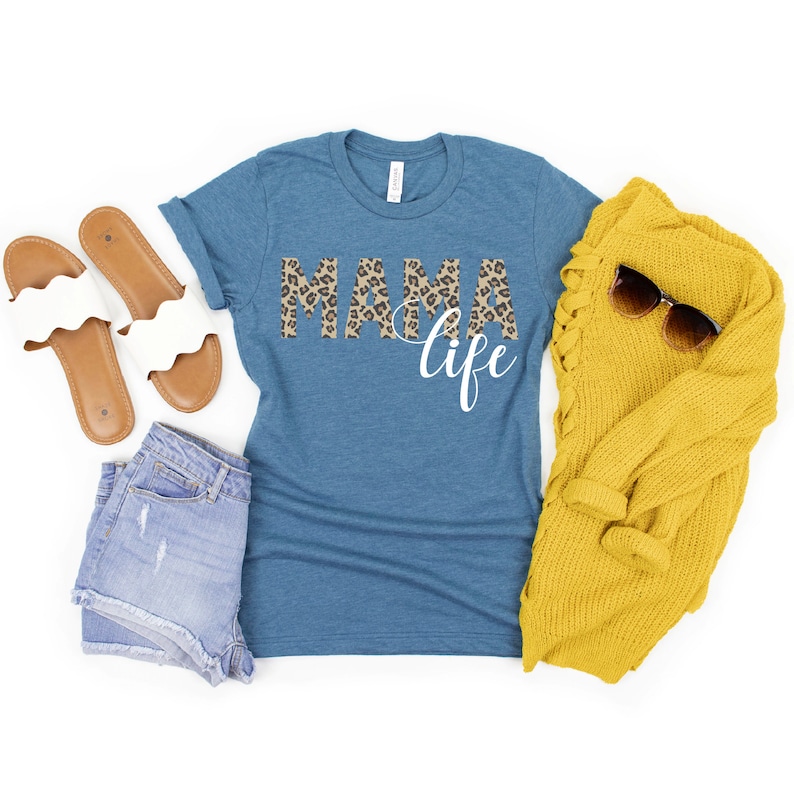 May include: A teal blue t-shirt with a leopard print "MAMA life" graphic. The shirt is paired with denim shorts, white sandals, and a yellow cardigan.