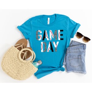 May include: A turquoise blue t-shirt with the text "GAME DAY" in white letters. The letters are decorated with a black and white leopard print and red baseball stitching.