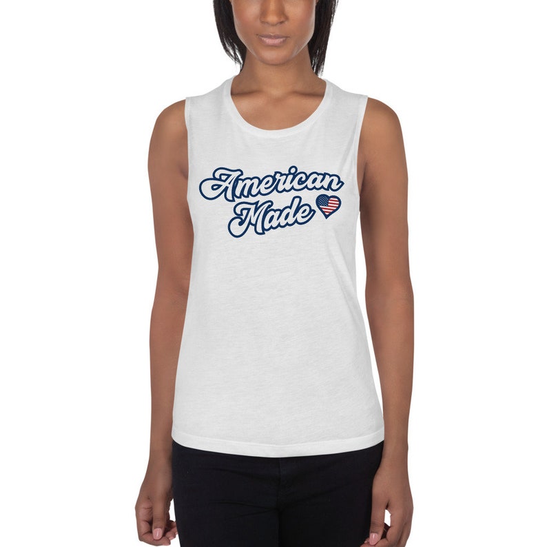 May include: White sleeveless muscle tank top with the text "American Made" in blue script with a small red, white, and blue heart with an American flag design.