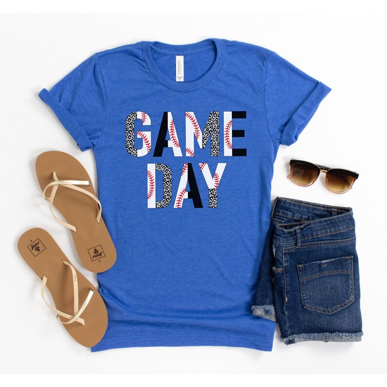 May include: A royal blue t-shirt with the words "GAME DAY" printed in black letters. The letters are made up of a baseball pattern with white and red stitching.