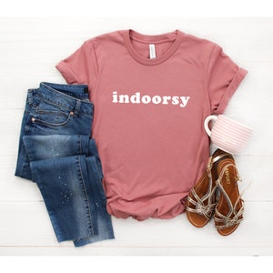 K&ouml;nnte beinhalten: Ein staubiges rosafarbenes T-Shirt mit dem Wort "indoorsy" in wei&szlig; auf der Vorderseite. Das Shirt wird mit einer blauen Jeans und braunen Sandalen kombiniert.
