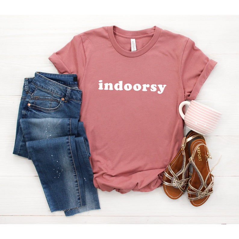 K&ouml;nnte beinhalten: Ein staubiges rosafarbenes T-Shirt mit dem Wort "indoorsy" in wei&szlig; auf der Vorderseite. Das Shirt wird mit einer blauen Jeans und braunen Sandalen kombiniert.