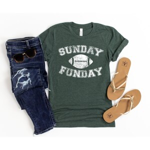 Op de afbeelding: Een groen T-shirt met een witte, verweerde afbeelding met de tekst "SUNDAY FUNDAY" met een voetbal in het midden.