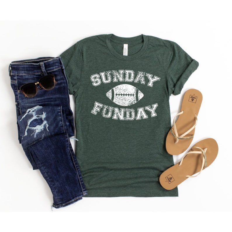 Op de afbeelding: Een groen T-shirt met een witte, verweerde afbeelding met de tekst "SUNDAY FUNDAY" met een voetbal in het midden.