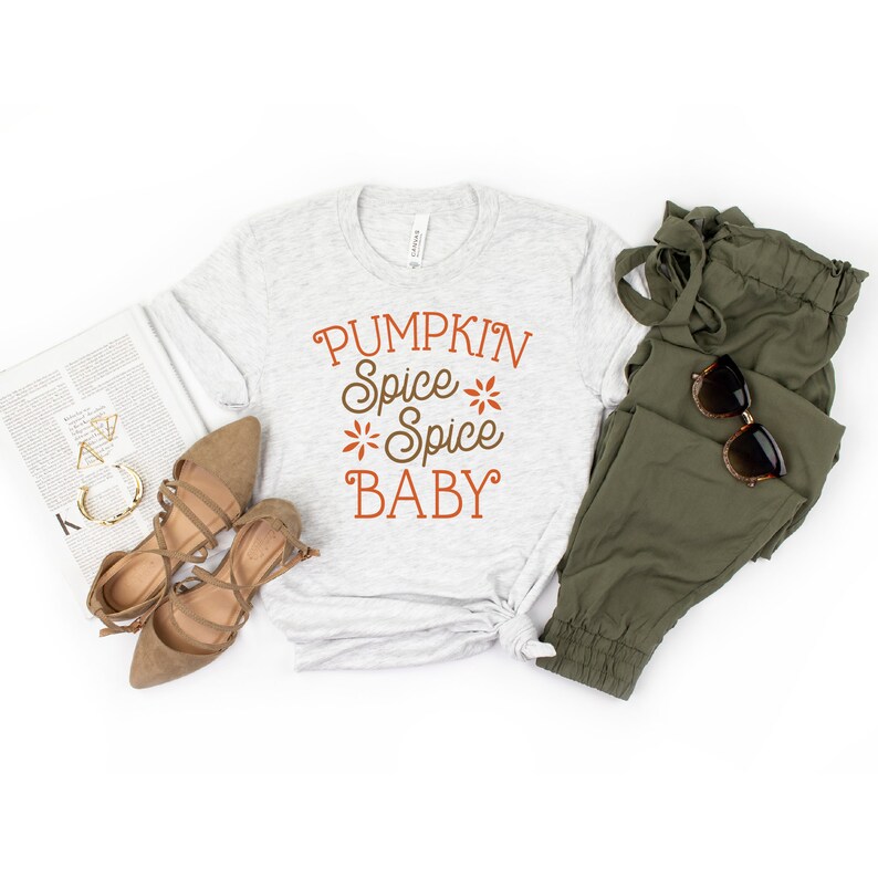 Op de afbeelding: Een lichtgrijs t-shirt met de tekst "PUMPKIN Spice Spice BABY" in oranje en bruin. Het shirt wordt gecombineerd met olijfgroene broeken, bruine sandalen, zonnebrillen en een tijdschrift op een witte ondergrond.