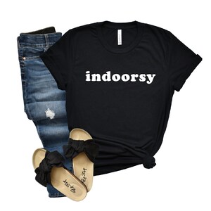 K&ouml;nnte beinhalten: Schwarzes T-Shirt mit dem Text "indoorsy" in wei&szlig;en Buchstaben. Das Shirt wird mit einer blauen Jeans und schwarzen Schleifensandalen kombiniert.